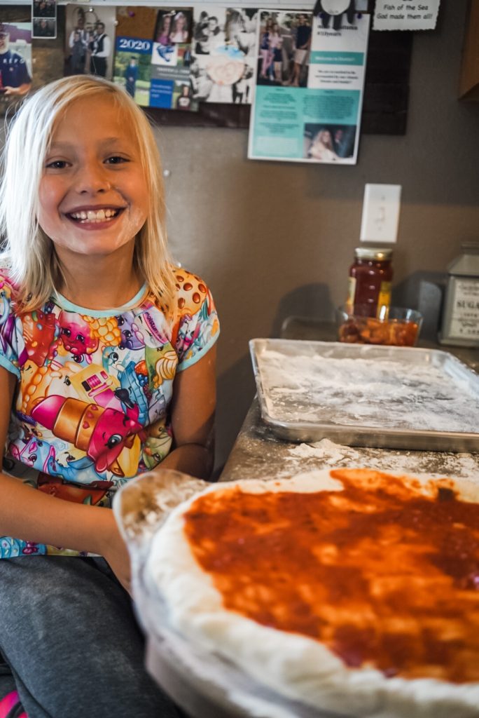 Girl smiling at camera with flour on her face sitting behind an unbaked pizza. 