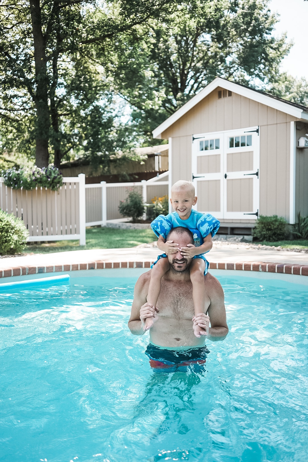 Small boy sitting on the shoulders of a man in a swimming pool. Boy is smiling at camera and has hands over man's eyes. 