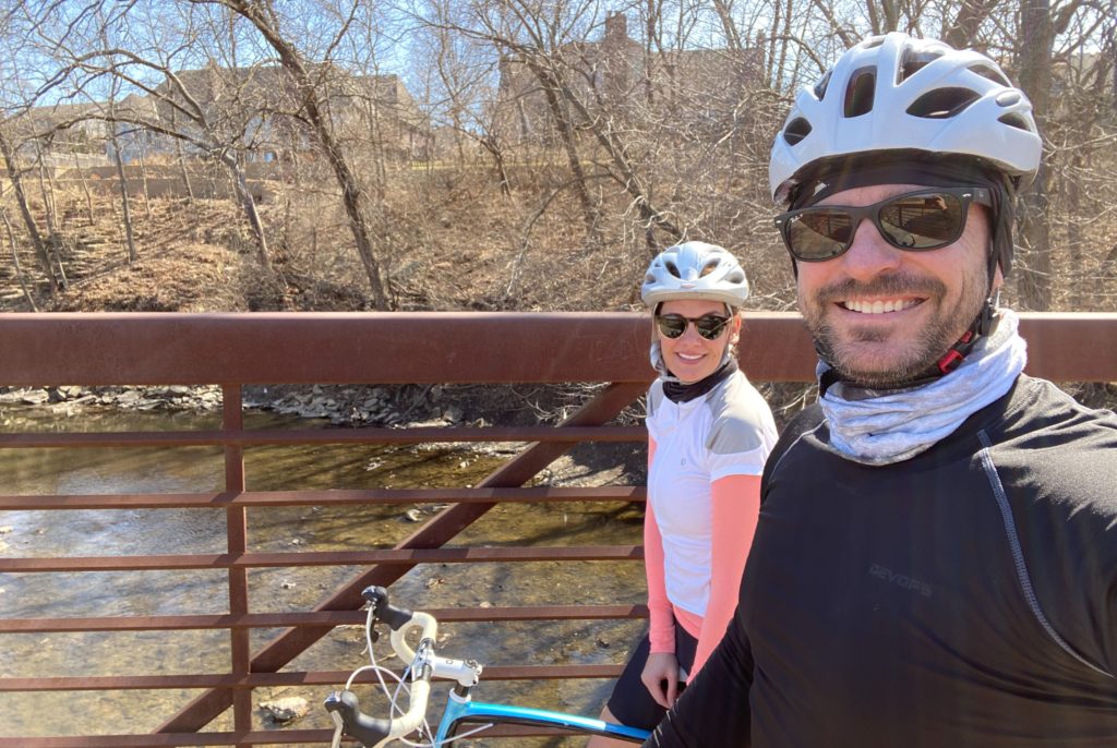 Man and woman smiling at camera sitting on bicycles with sunglasses on and bike helmets. In background there is a creek. 