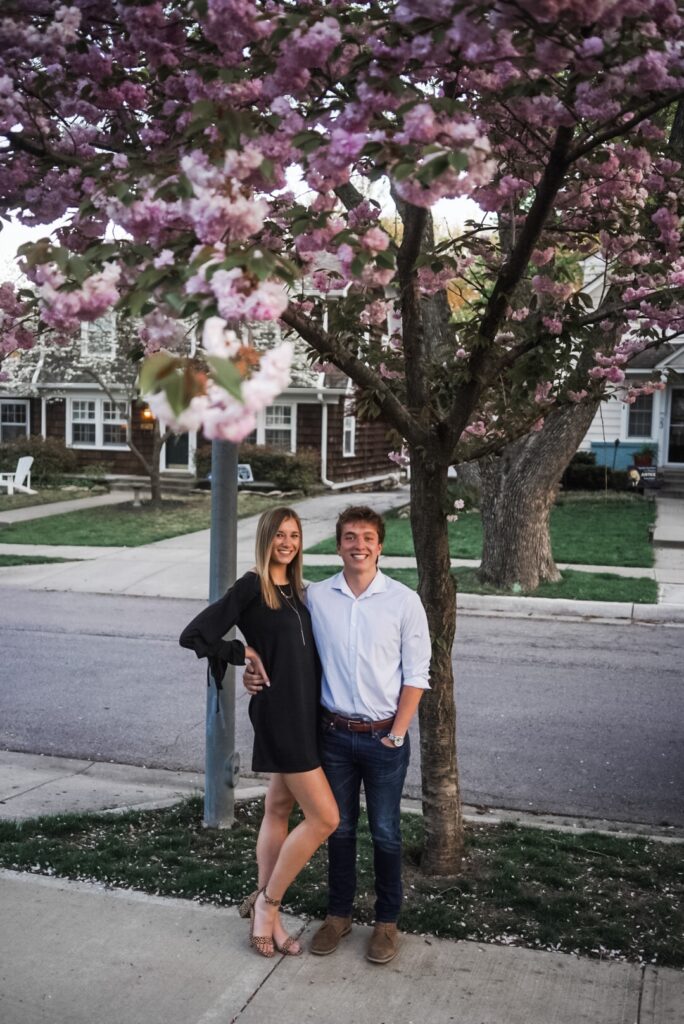 White girl with blonde hair wearing black dress standing next to white boy with light blue shirt. Both are standing underneath a tree with pink blossoms.